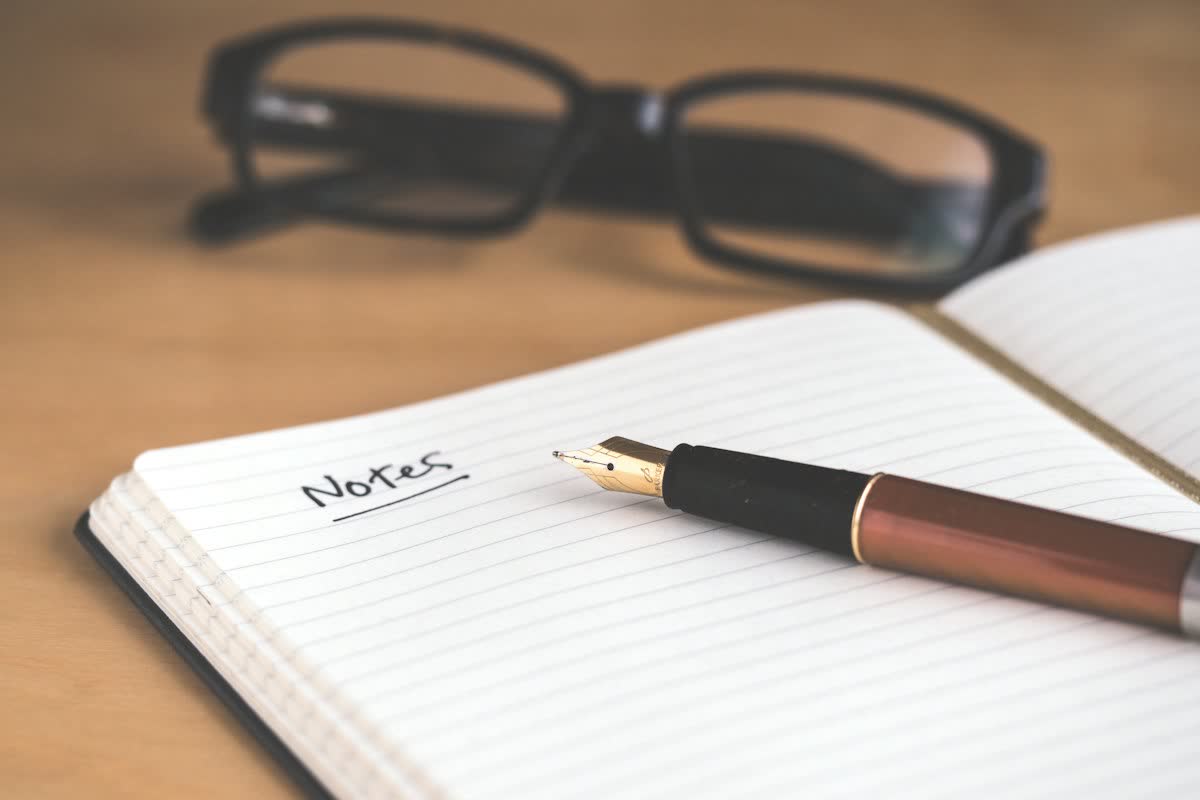 Buku catatan bergaris bertuliskan Notes dengan pulpen dan kacamata di atas meja kayu
