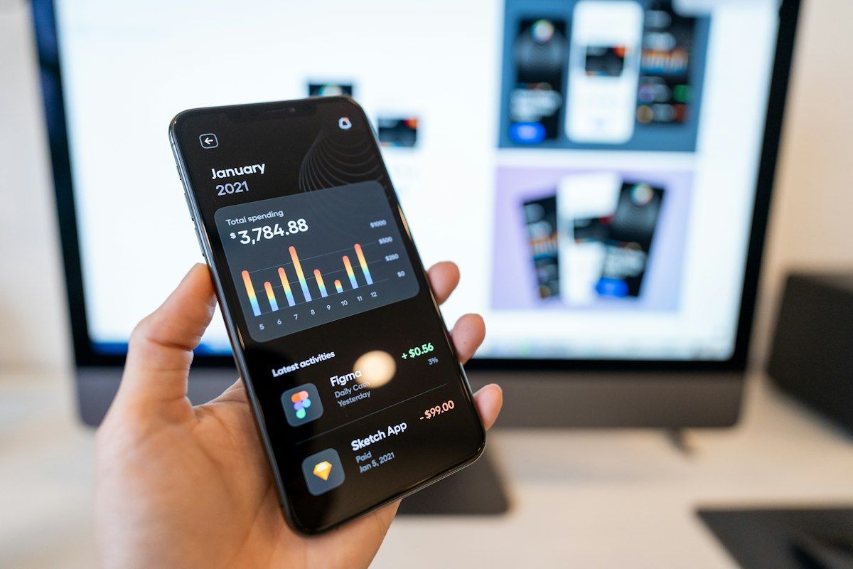 Hand holding a phone showing a finance app, with app designs on a monitor behind it