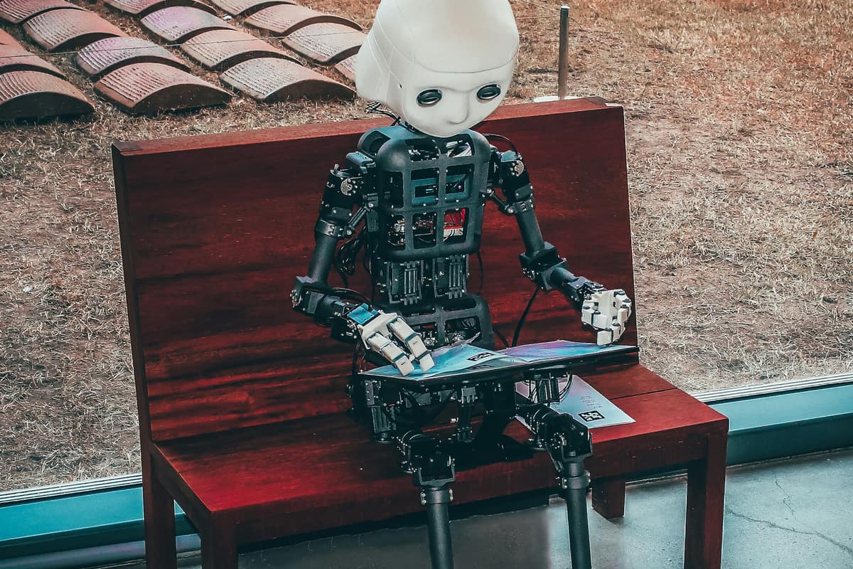 Robot humanoid kecil duduk di bangku sambil membaca laptop