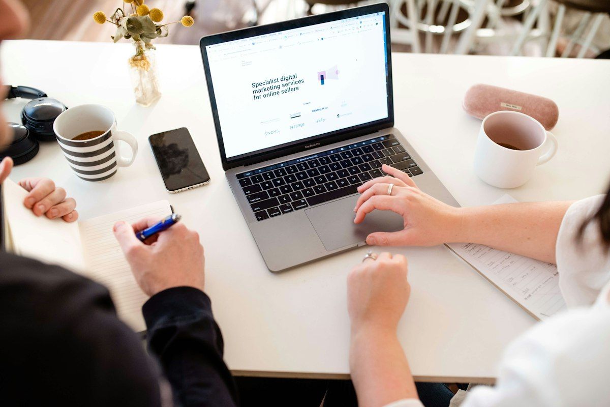 Two people planning a website at a desk, with a laptop showing a homepage, notes, and coffee cups